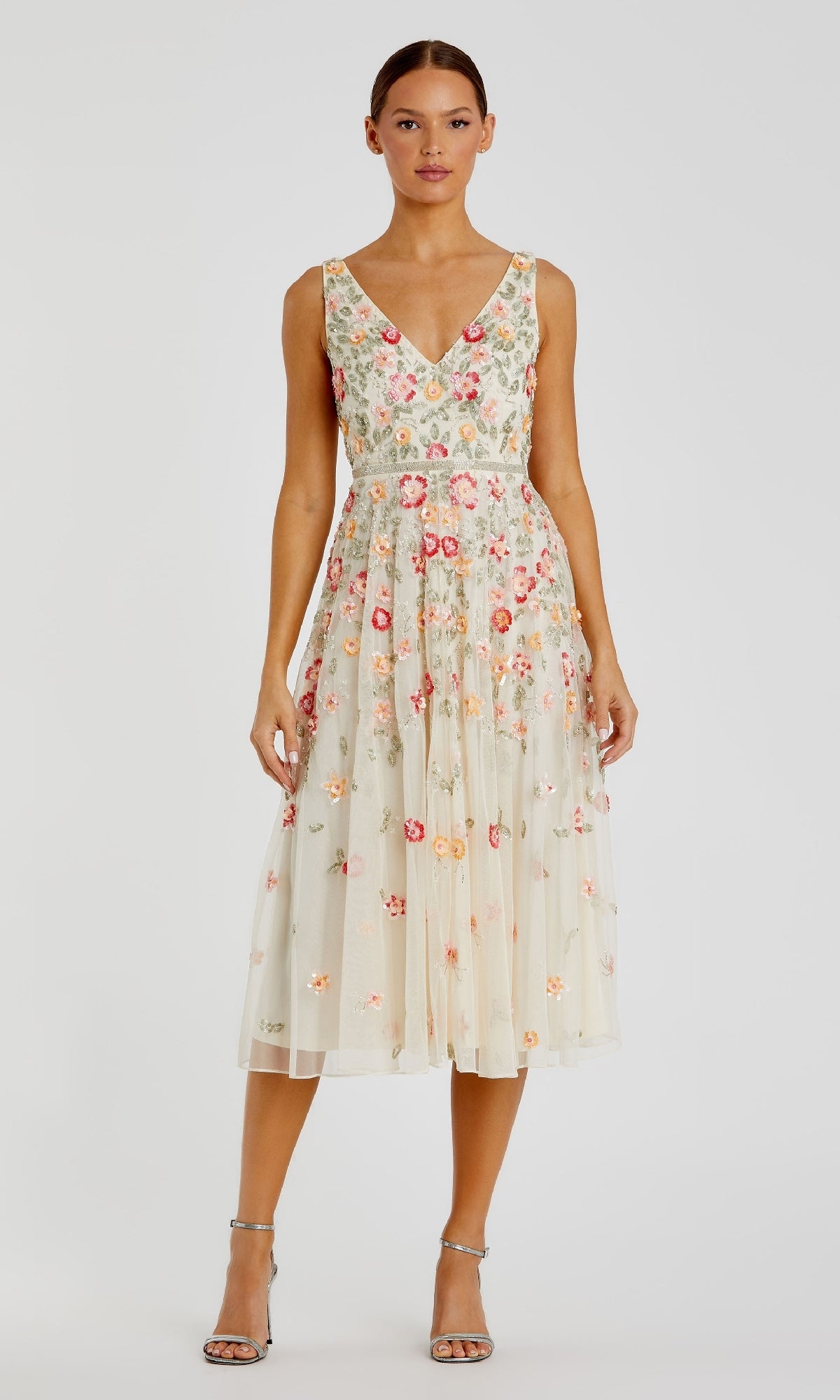 A woman stands facing forward in the Mac Duggal 5527 Floral-Embroidered Party Dress—a sleeveless, V-neck, cream tea-length dress with colorful floral embroidery—paired with silver high-heeled sandals against a plain background.